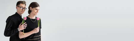 Young gay in eyeglasses holding tulip near smiling boyfriend isolated on grey, bannerの写真素材