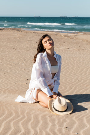 young woman in white shirt and swimwear holding straw hat and sitting on golden sand near blue oceanの写真素材