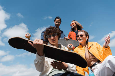 low angle view of young man holding skateboard near happy interracial friends outdoorsの写真素材