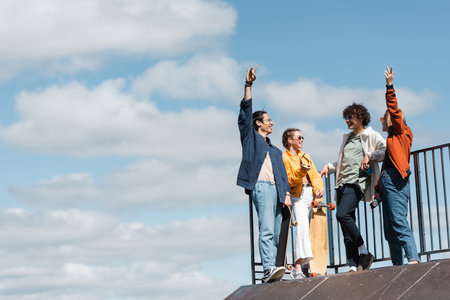 cheerful interracial friends gesturing on skate ramp under blue and cloudy skyの写真素材