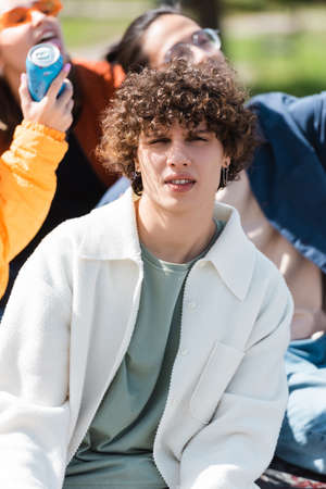young and curly man looking at camera near interracial friends on blurred background