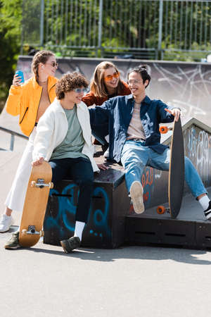 cheerful and trendy multiethnic friends sitting on ledge in skate parkの写真素材