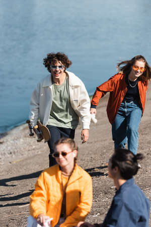 cheerful man with skateboard running on river bank near multicultural friends
