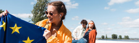 joyful woman in stylish sunglasses holding european union near friends on blurred background, bannerの写真素材
