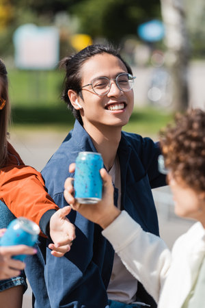 young asian man in eyeglasses and earphone smiling near blurred friends with soda cansの写真素材
