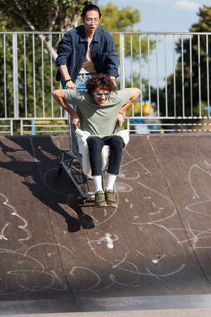 cheerful interracial friends having fun with shopping cart on ramp in skate parkの写真素材