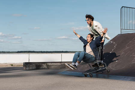 excited interracial friends moving on shopping cart down from skate ramp