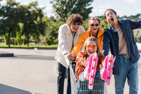 woman in rollers skates riding in shopping cart near interracial friends outdoorsの写真素材