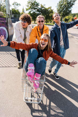 thrilled woman with outstretched hands riding in shopping trolley near interracial friendsの写真素材
