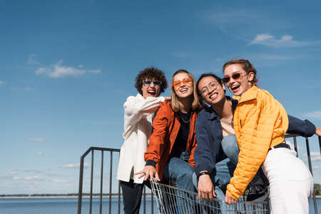 excited multicultural friends with shopping trolley laughing at camera against blue skyの写真素材