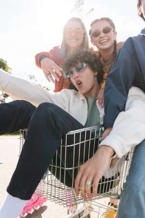 astonished man riding in shopping cart near cheerful friendsの写真素材