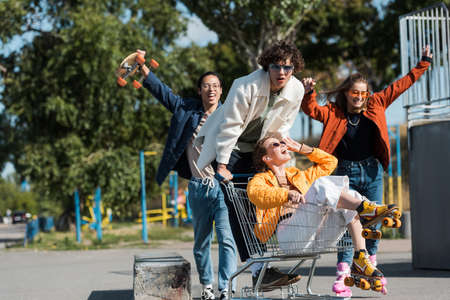 young and cheerful multiethnic skaters having fun with shopping cart in park