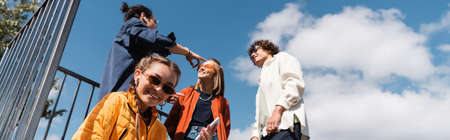 low angle view of young woman smiling at camera near interracial friends against cloudy sky, bannerの写真素材