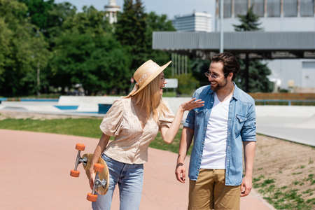 Cheerful woman in sun hat holding longboard and talking to boyfriend in skate park