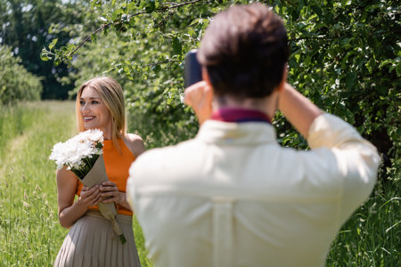 Positive woman with bouquet posing near blurred boyfriend with retro camera in park
