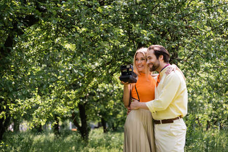 Smiling man hugging stylish girlfriend with vintage camera in summer parkの写真素材