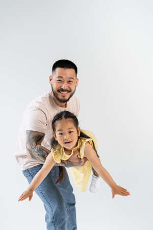 cheerful asian father holding in arms joyful preschooler daughter in yellow dress isolated on greyの写真素材