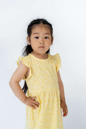 brunette preschooler asian girl posing with hand on hip while looking at camera isolated on grey