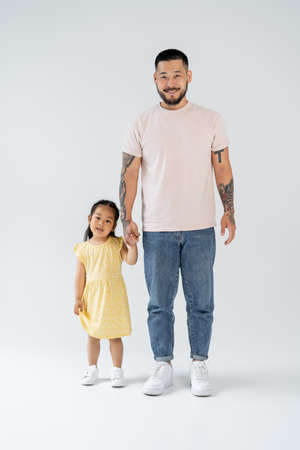 full length of happy and tattooed asian father holding hands with preschooler daughter on greyの写真素材