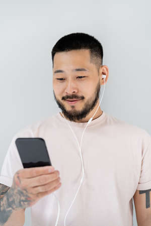 cheerful asian man in wired earphones listening music and holding smartphone isolated on grey