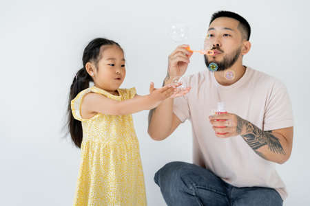 asian father blowing soap bubbles near amazed preschooler daughter isolated on greyの写真素材