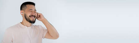 happy and tattooed asian man with closed eyes wearing wired earphones isolated on grey, banner