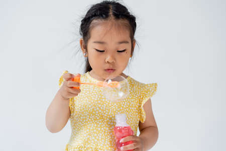 preschooler asian girl in dress holding bottle with soap bubbles isolated on grey