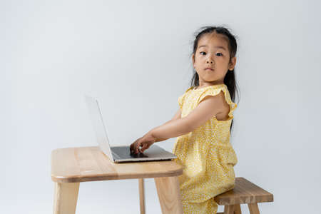 asian preschooler girl in yellow dress using laptop and looking at camera isolated on grey