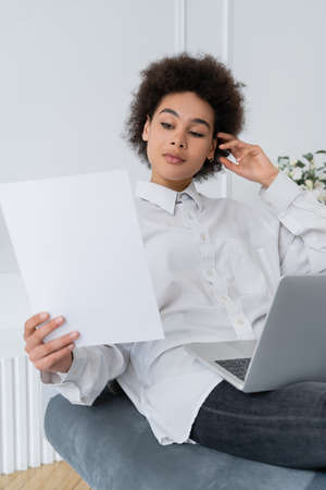 african american woman looking at blank paper while using laptop at home