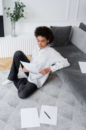 high angle view of african american woman holding blank document while sitting on carpet near grey...