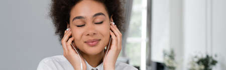 happy african american woman with closed eyes listening music in wired earphones, banner