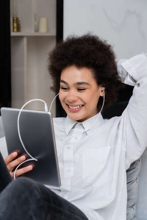 joyful african american woman in wired earphones holding digital tablet while watching comedy movieの写真素材
