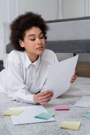 curly african american woman holding blank document near papers on carpet