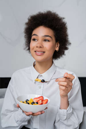 happy african american woman eating corn flakes with fresh berries at homeの写真素材