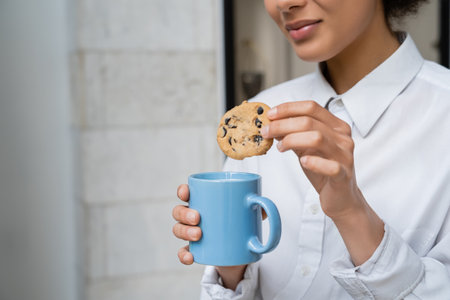 cropped view of happy african american woman holding cup of milk and cookie with chocolate chips