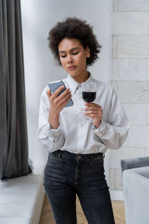 sad african american woman holding glass of red wine and looking at smartphoneの写真素材
