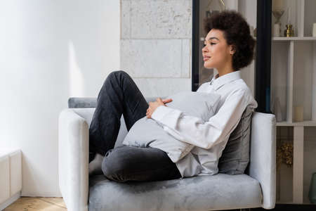 dreamy and curly african american woman in white shirt with collar sitting with pillow on grey armchairの写真素材