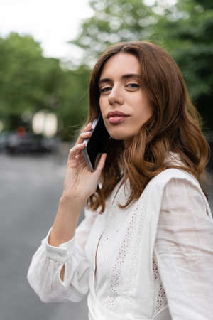 Brunette woman talking on smartphone and looking at camera on street in Parisの写真素材