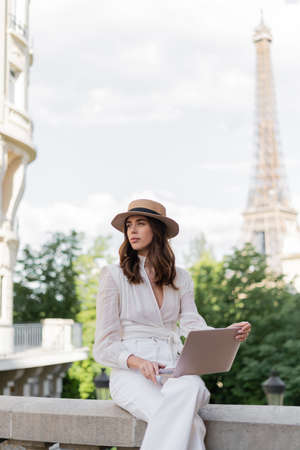 Fashionable freelancer in sun hat using laptop on urban street with blurred Eiffel tower at background in Parisの写真素材