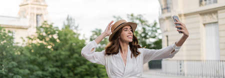 Cheerful young woman in sun hat taking selfie on smartphone with blurred Eiffel tower at background in France, bannerの写真素材