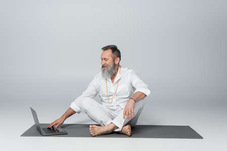 smiling spiritual mentor sitting on yoga mat in easy pose near laptop on grey