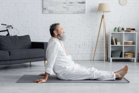 side view of bearded guru man sitting on yoga mat while meditating at homeの写真素材