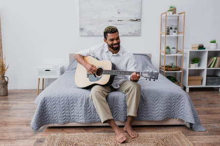 barefoot and happy african american man with dyed hair and beard playing acoustic guitar in bedroom