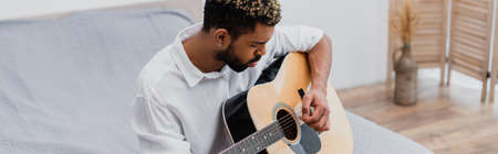 bearded young african american man with dyed hair playing acoustic guitar in bedroom, bannerの写真素材