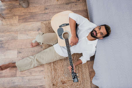 top view of pleased african american man with dyed hair holding acoustic guitar and leaning on bed