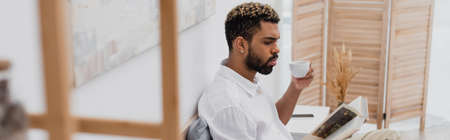 young african american man with dyed hair reading book and holding cup of coffee in modern bedroom,...