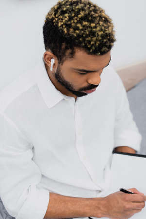 high angle view of young african american man in wireless earphone writing in notebookの写真素材