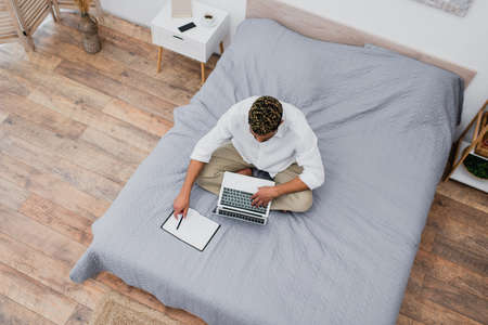 top view of young african american man with dyed hair using laptop while sitting on bed