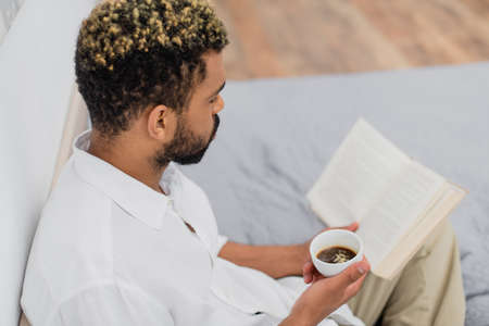 high angle view of young african american man with dyed hair reading novel and holding cup of coffee...