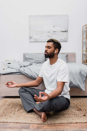 bearded african american man in wireless earphone meditating with closed eyes while sitting near bed at homeの写真素材